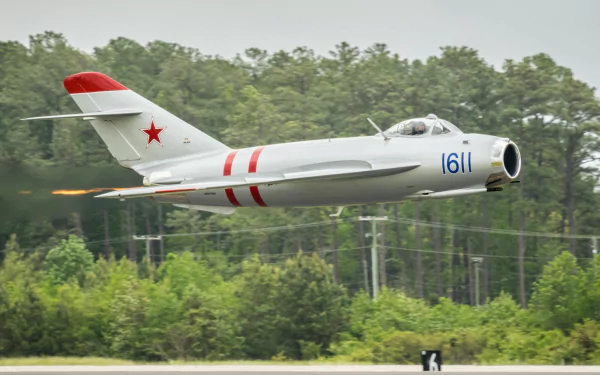 HD PC desktop wallpaper: military Mikoyan-Gourevitch MiG-17 jet, white with red markings and tail star, flying low above runway with dense green forest backdrop.