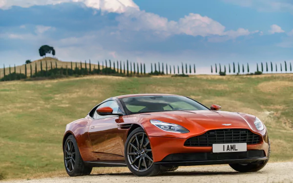 Orange Aston Martin DB11 grand tourer parked on a dirt road with rolling hills and a partly cloudy sky in the background, presented as a 4K Ultra HD desktop wallpaper.