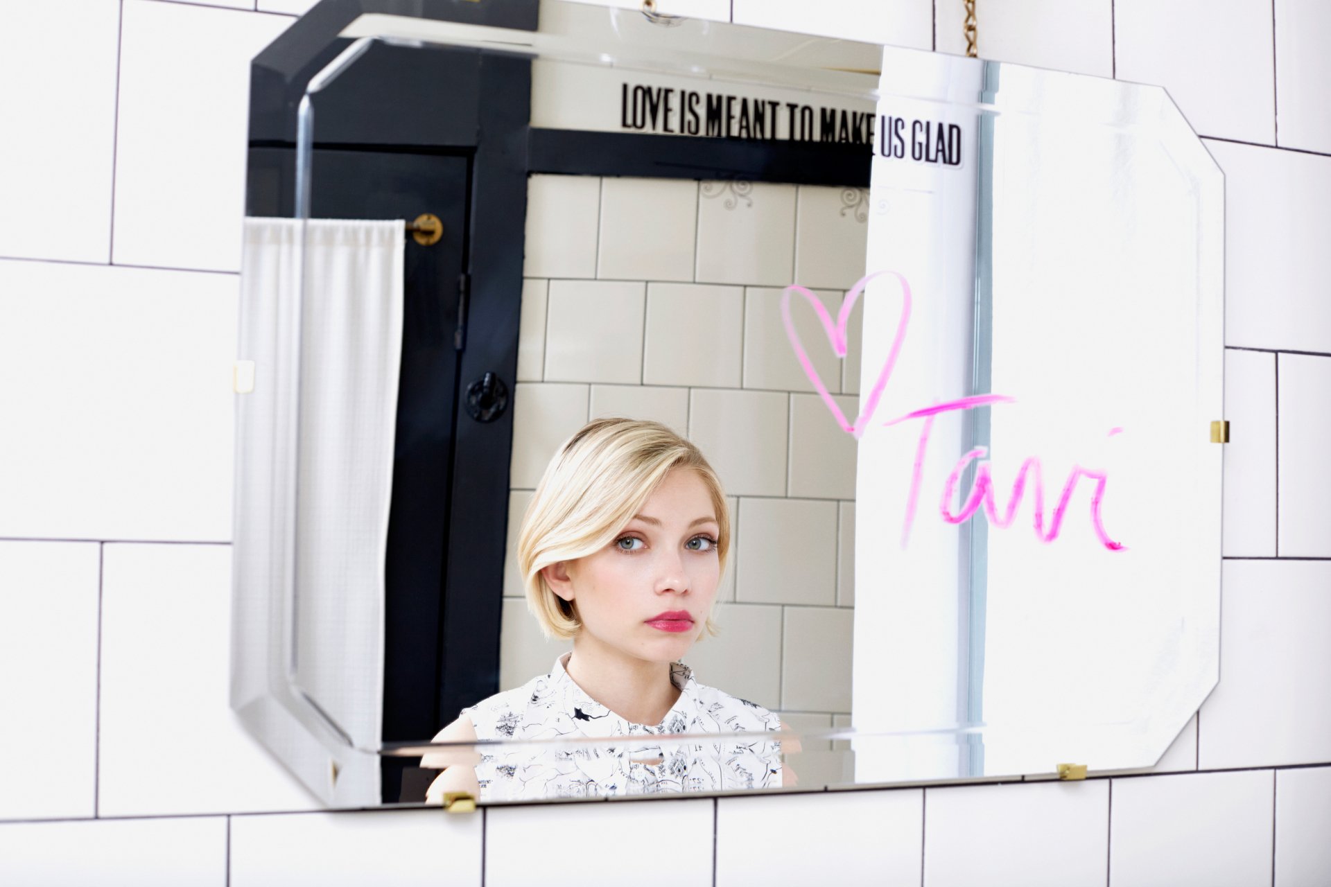 HD desktop wallpaper showing a young blonde woman's face reflected in a tiled bathroom mirror, wearing pale lipstick with a pink handwritten signature on the glass.