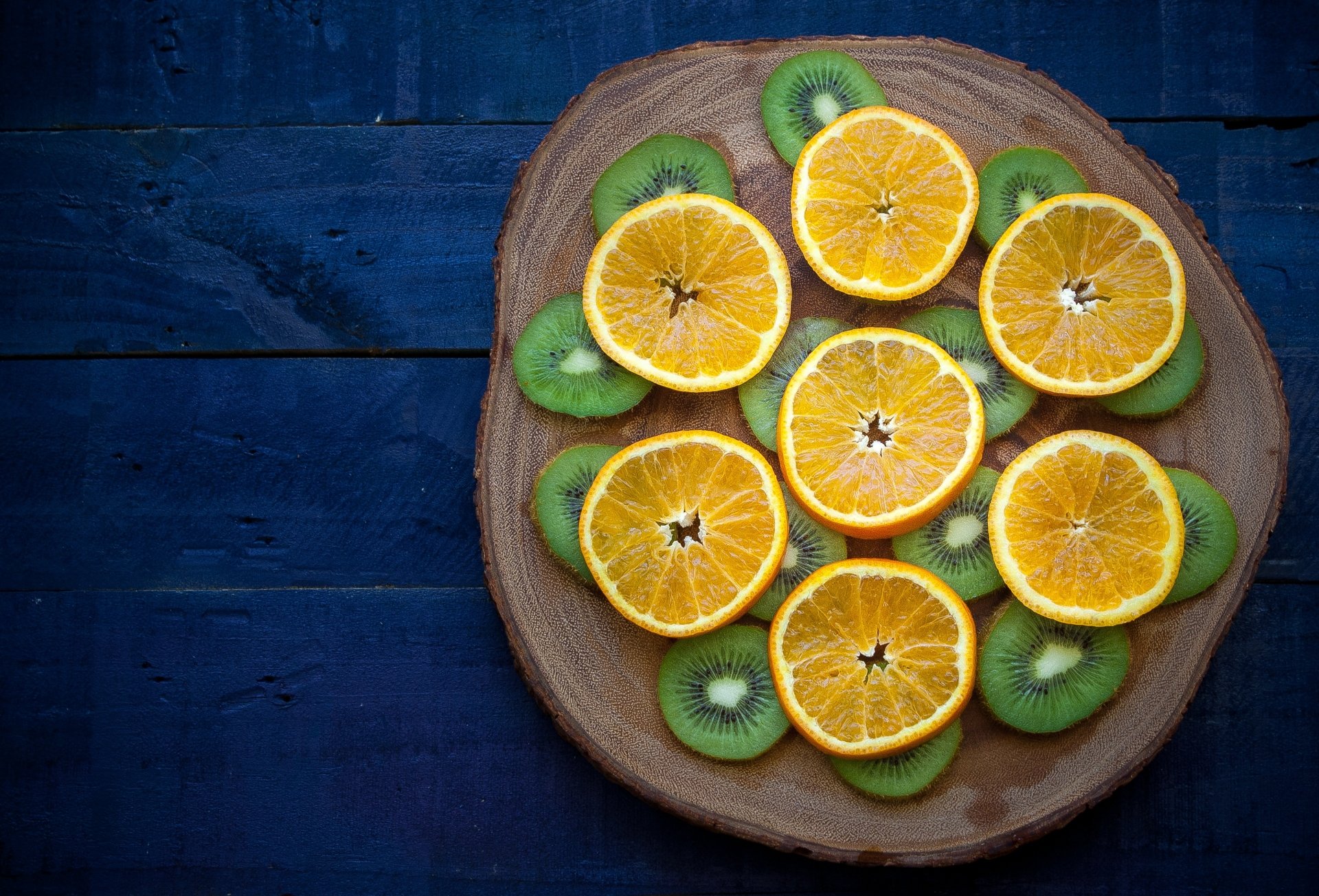 HD desktop wallpaper featuring a wooden plate with sliced orange fruits and kiwi arranged on a dark blue wooden background.