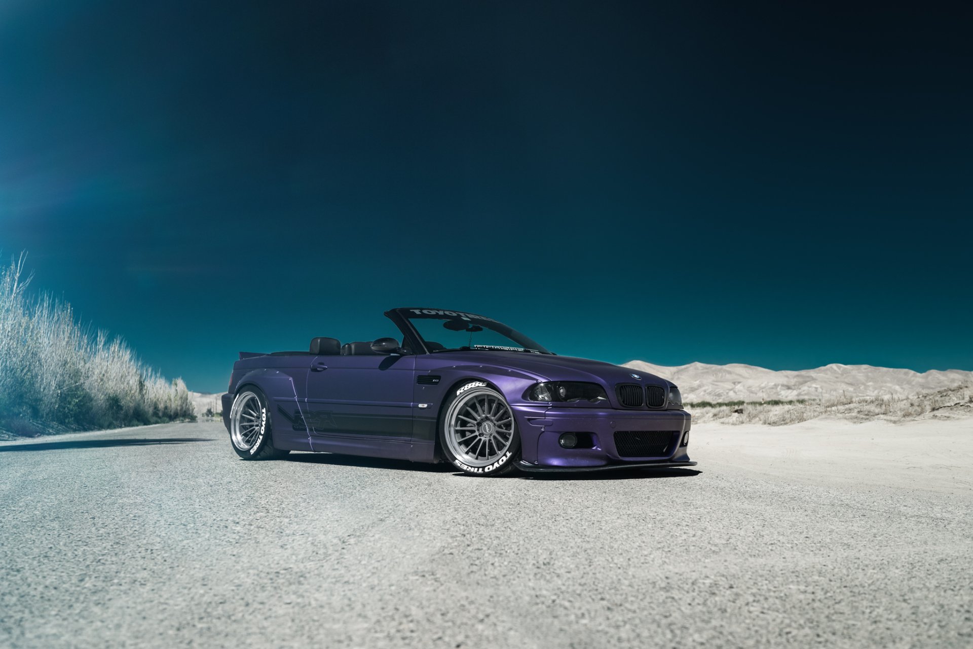 HD desktop wallpaper featuring a sleek purple BMW M3 convertible parked on a sandy terrain under a clear blue sky.