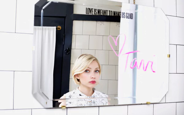 HD desktop wallpaper showing a young blonde woman's face reflected in a tiled bathroom mirror, wearing pale lipstick with a pink handwritten signature on the glass.