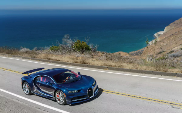 A stunning Bugatti Chiron supercar drives along a scenic coastal road, with a clear blue ocean in the background, creating an impressive HD wallpaper for any car enthusiast.