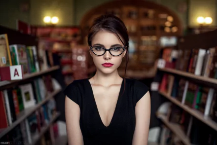 A woman wearing glasses stands confidently in a library, with bookshelves on either side. This HD desktop wallpaper features Oktyabrina Maximova and serves as a striking background image.