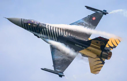 A General Dynamics F-16 Fighting Falcon jet fighter soaring through the sky, trailing smoke against a blue backdrop, showcasing its military prowess and sleek design.