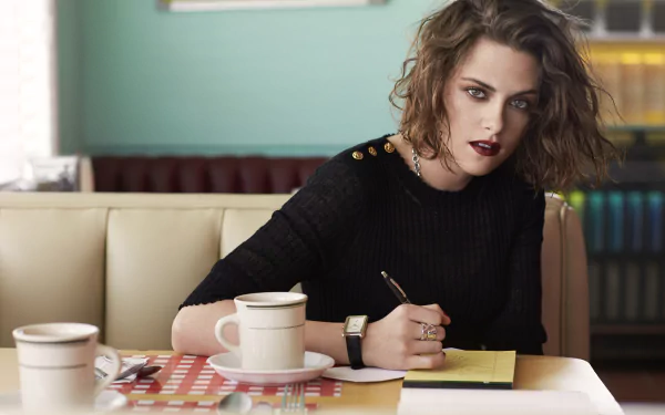 A stylish Kristen Stewart sits at a café table, writing on a yellow notepad, with coffee cups beside her, captured in vivid 4K Ultra HD quality.