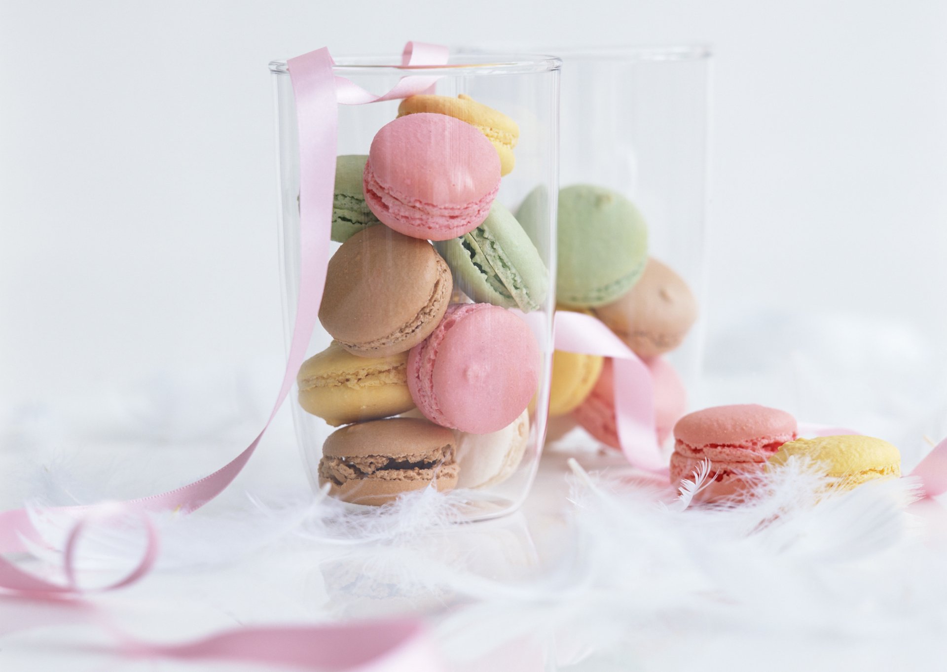 A vibrant display of colorful macarons in a clear glass container, surrounded by soft white feathers and pink ribbon strands, creating a delightful and whimsical food-themed wallpaper.