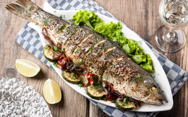 A beautifully seasoned grilled whole fish served on a bed of lettuce with roasted vegetables, lemon wedges, and a glass of white wine, presented in 4K Ultra HD.