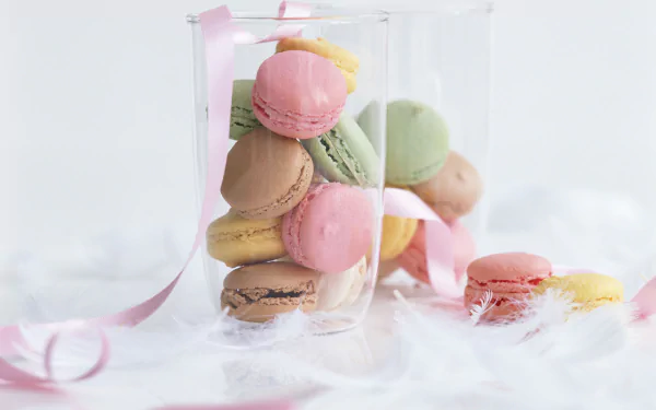 A vibrant display of colorful macarons in a clear glass container, surrounded by soft white feathers and pink ribbon strands, creating a delightful and whimsical food-themed wallpaper.