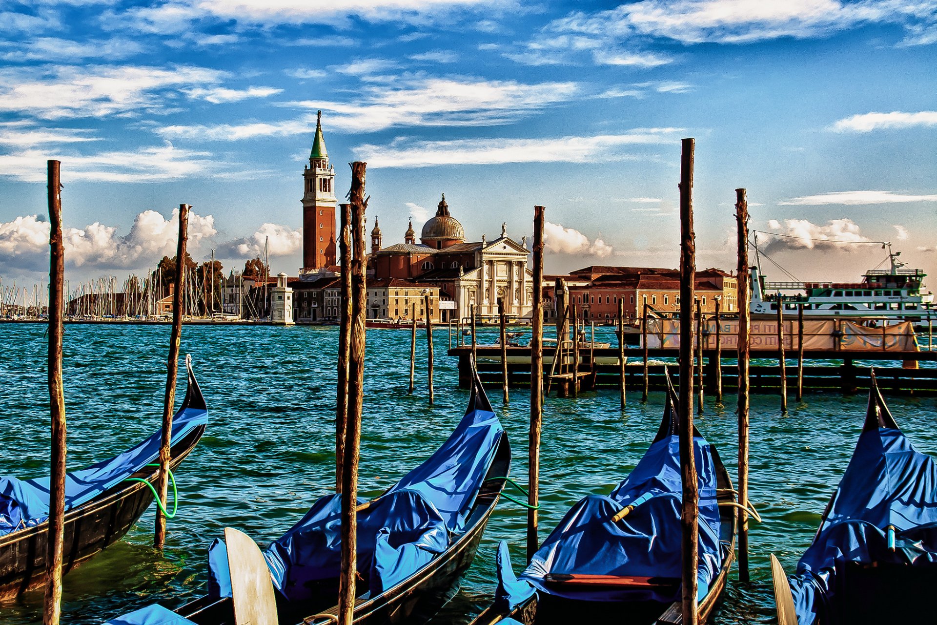 A vibrant HD desktop wallpaper of Venice, Italy, showcasing gondolas on a canal with historic city buildings and a bright blue sky in the background.