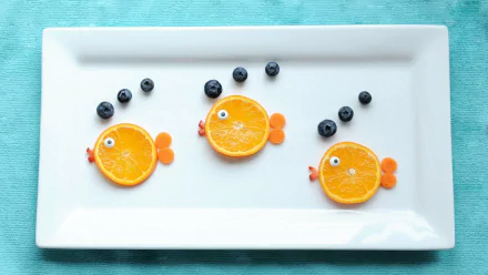 A creative still life HD desktop wallpaper showing three orange slice fish with blueberry eyes and bubbles on a white plate against a light background.