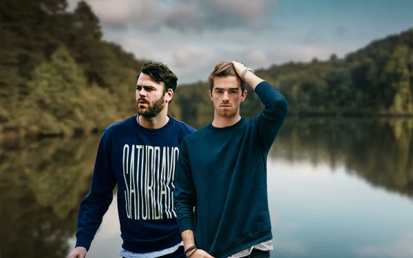 HD desktop wallpaper featuring The Chainsmokers standing by a serene lake and forest background. Both are wearing casual sweatshirts, with one of them running a hand through his hair.