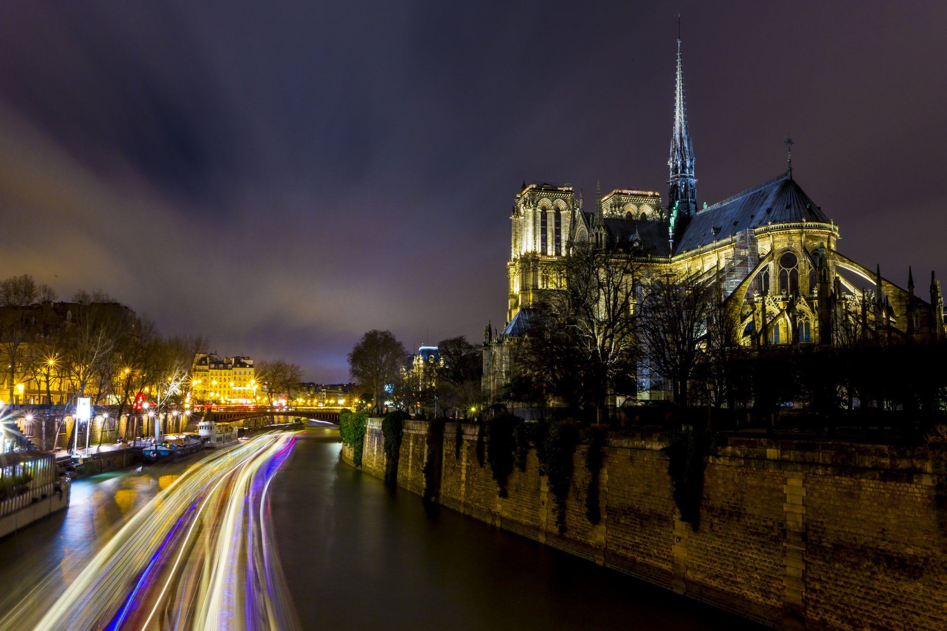 Nighttime light trails create a vibrant time-lapse effect along the Seine beside the illuminated Notre-Dame de Paris cathedral, captured in HD for a striking desktop wallpaper.