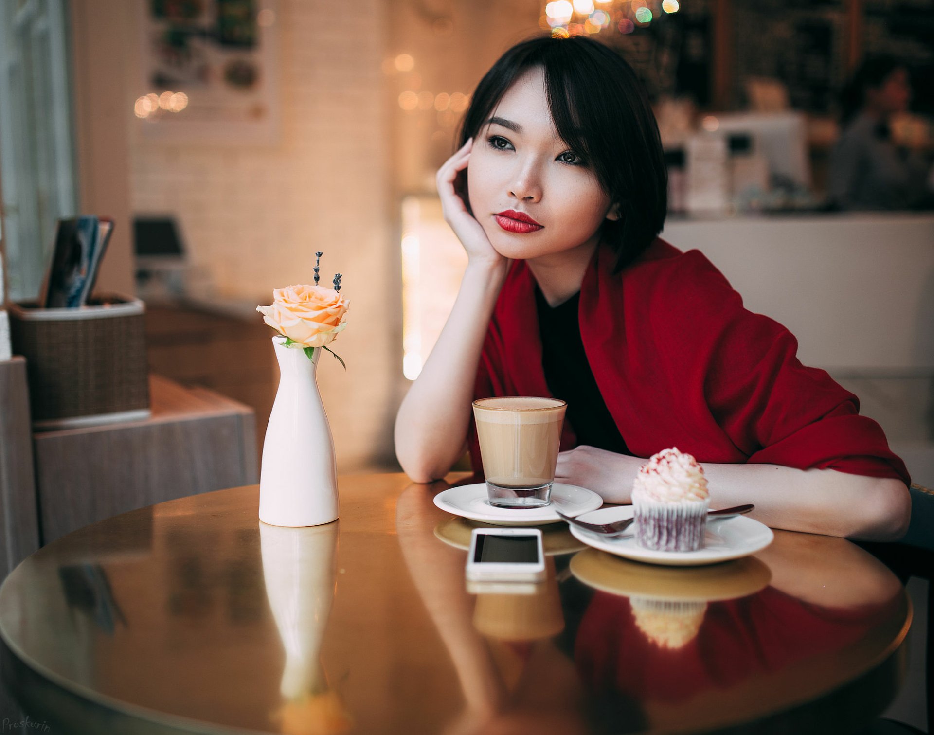 A brunette woman with brown eyes and red lipstick rests her head on her hand at a café table with a coffee, cupcake, and flower vase, captured in HD desktop wallpaper style.