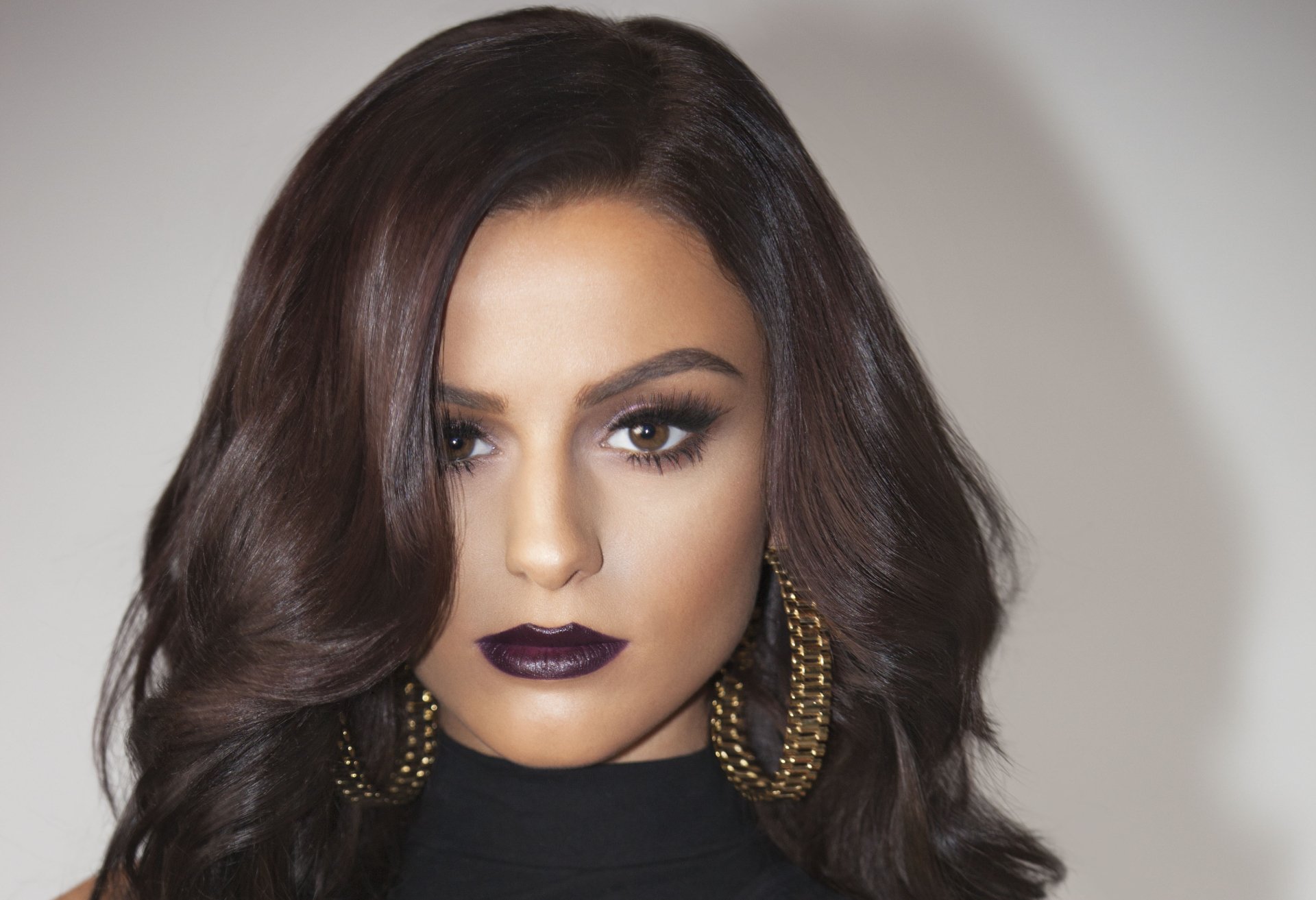 Close-up portrait of English singer Cher Lloyd with brunette hair, brown eyes, dark lipstick, and large hoop earrings, set against a plain background.
