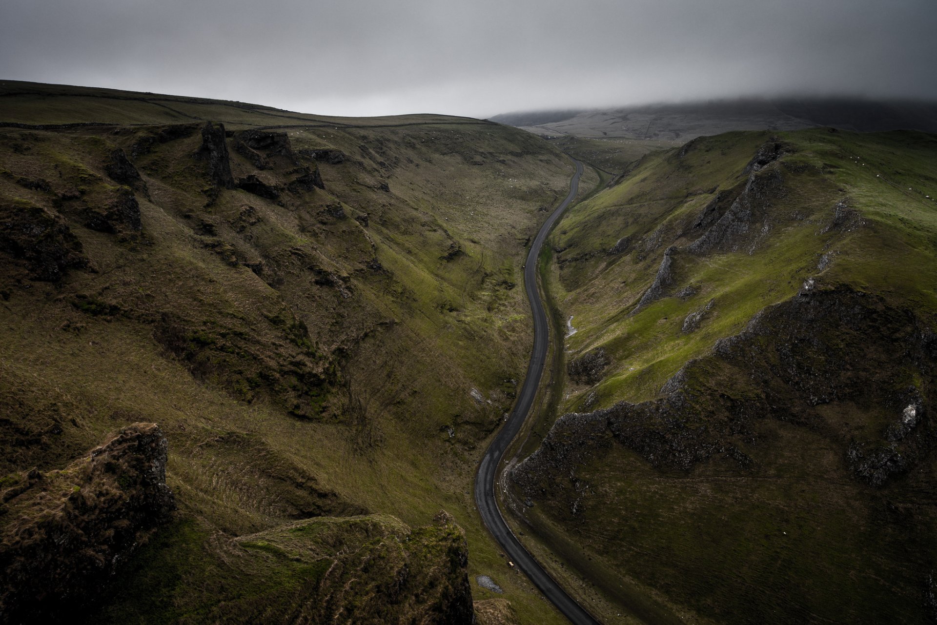 HD PC desktop wallpaper of a winding man-made road cutting through a misty green mountain landscape under an overcast sky.