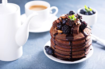 A stack of chocolate pancakes topped with blackberries, blueberries, and nuts, drizzled with chocolate sauce, served with coffee and a teapot in the background.