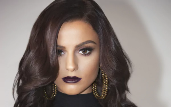 Close-up portrait of English singer Cher Lloyd with brunette hair, brown eyes, dark lipstick, and large hoop earrings, set against a plain background.