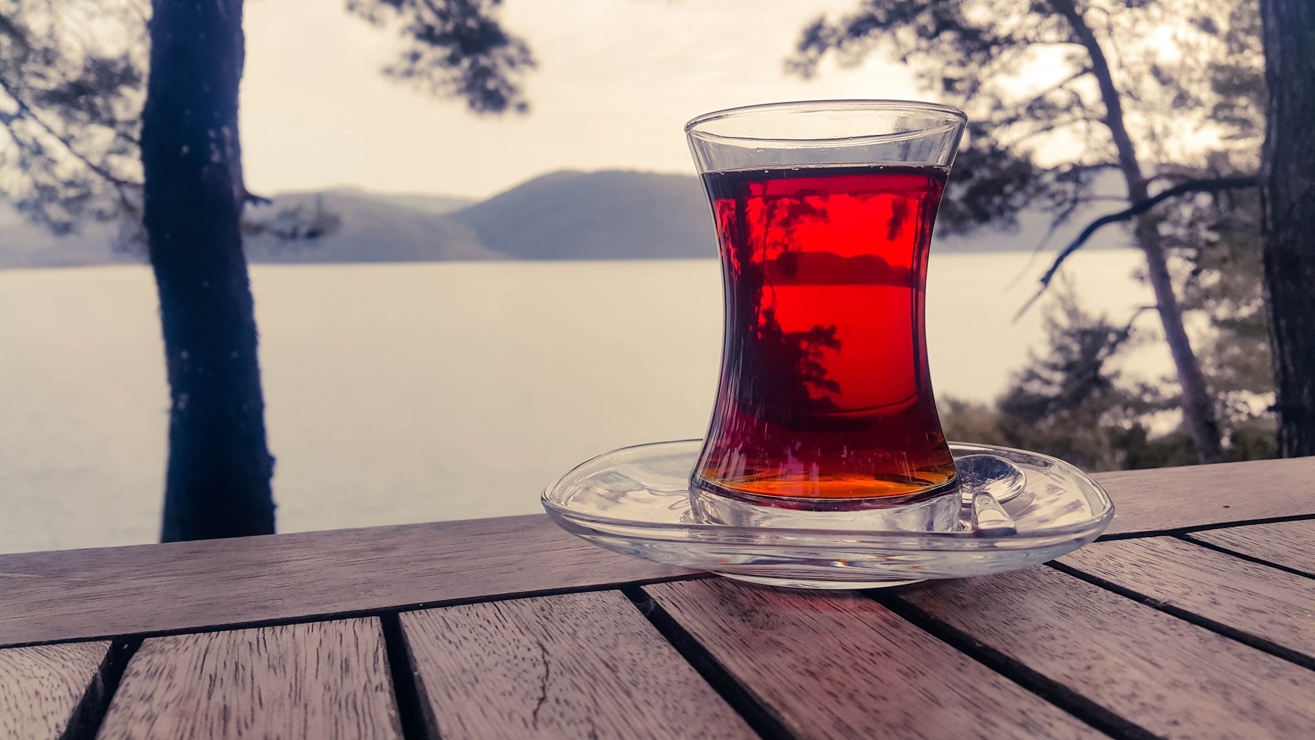 A glass of deep red tea on a wooden table overlooking a serene lake and trees, captured in stunning 4K Ultra HD for PC desktop wallpaper and background.