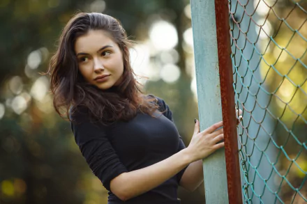 A brunette woman with brown eyes stands next to a fence, with a soft bokeh background. This HD desktop wallpaper captures her looking away thoughtfully.
