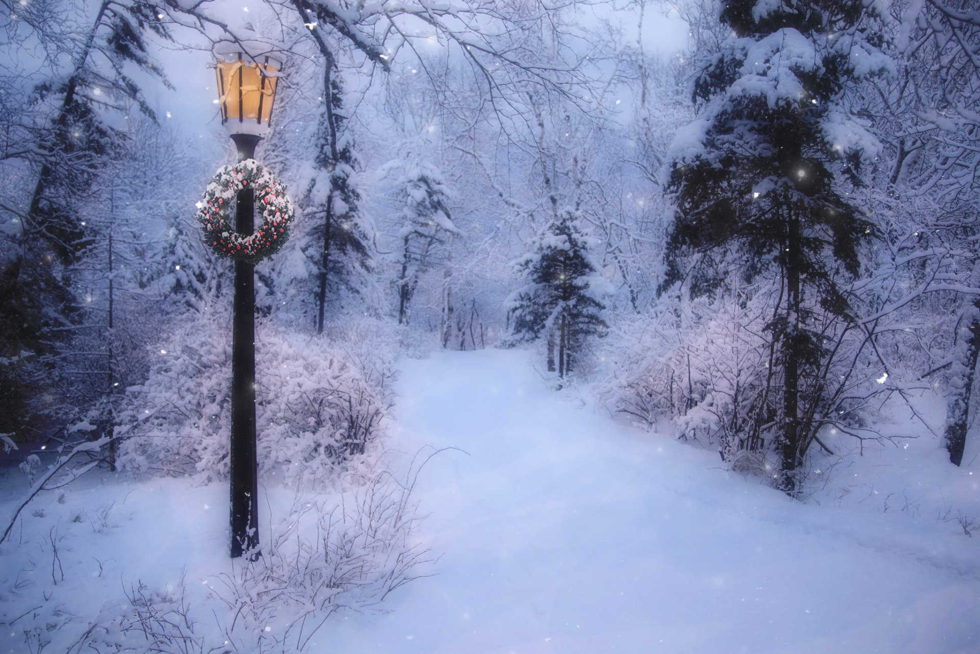 A snow-covered road winds through a winter landscape adorned with a festive wreath on a lamppost, capturing the serene beauty of Christmas in a snowfall.