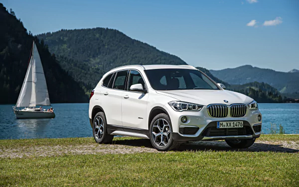 A white BMW X1 SUV parked by a lakeside with mountains in the background. 4K Ultra HD PC desktop wallpaper capturing the vehicle and serene nature scene.