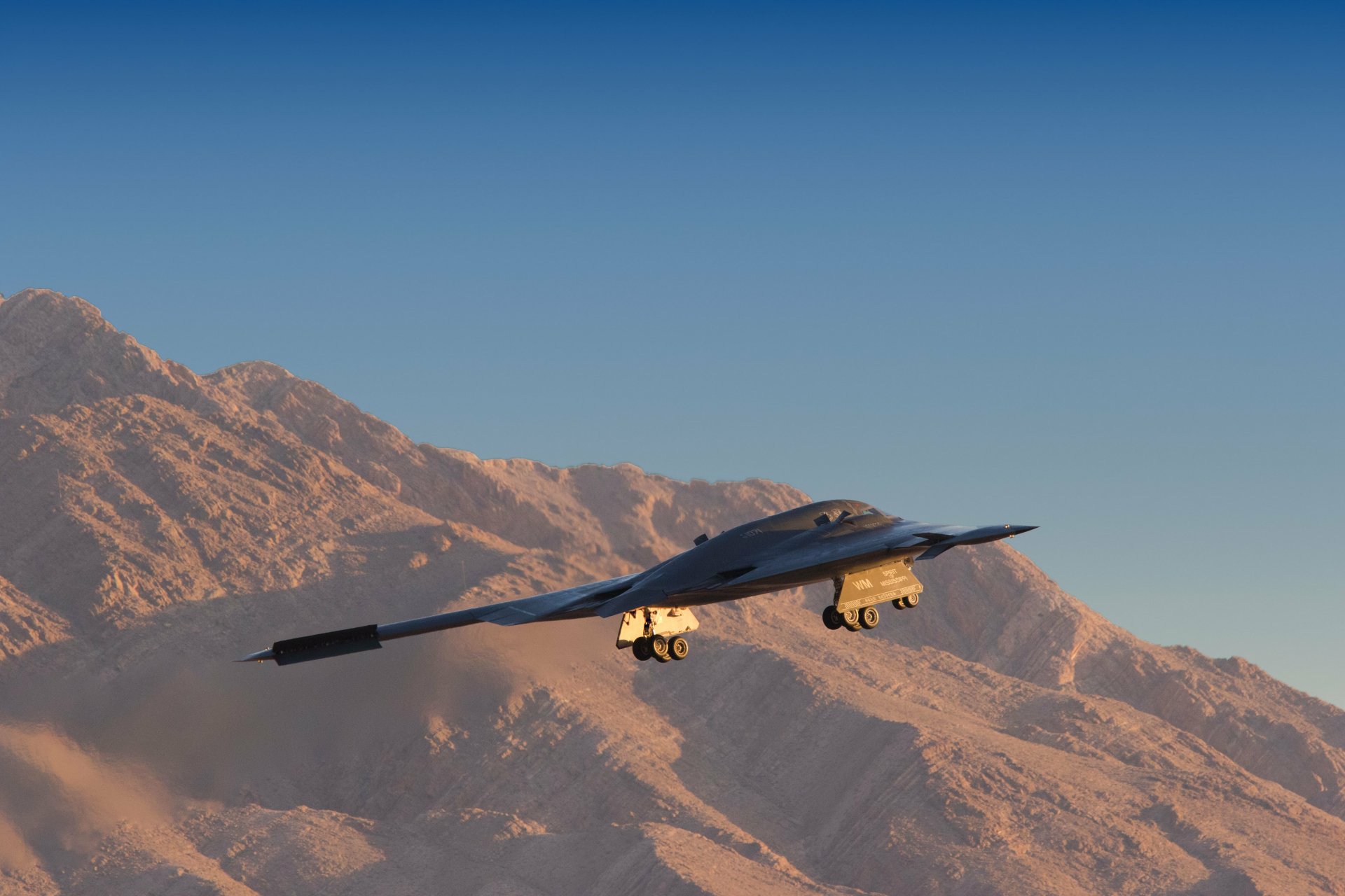 Northrop B-2 Spirit military stealth bomber flying over rugged desert mountains beneath a clear blue sky — 5K Ultra HD PC desktop wallpaper.