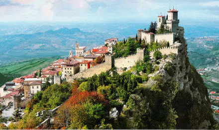 Panoramic HD desktop wallpaper of the Fortress of Guaita perched on a mountainous hill, surrounded by a village with houses and lush greenery.