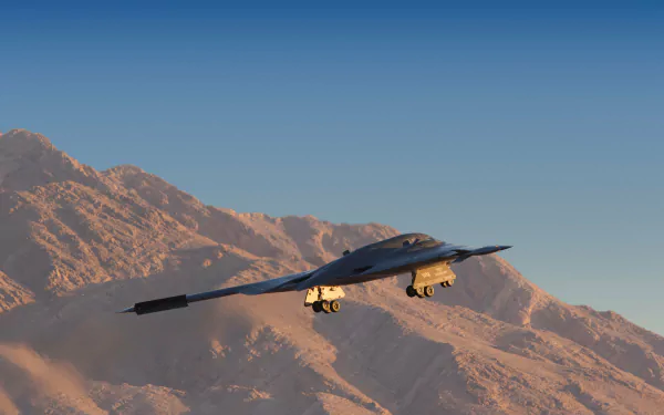 Northrop B-2 Spirit military stealth bomber flying over rugged desert mountains beneath a clear blue sky — 5K Ultra HD PC desktop wallpaper.