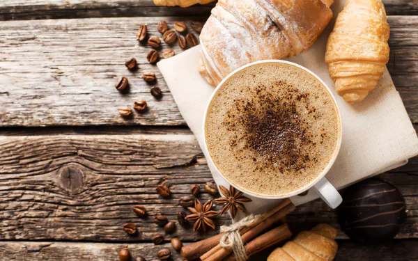 4K Ultra HD wallpaper featuring a coffee cup sprinkled with cinnamon, surrounded by croissants, star anise, and coffee beans on a rustic wooden surface.