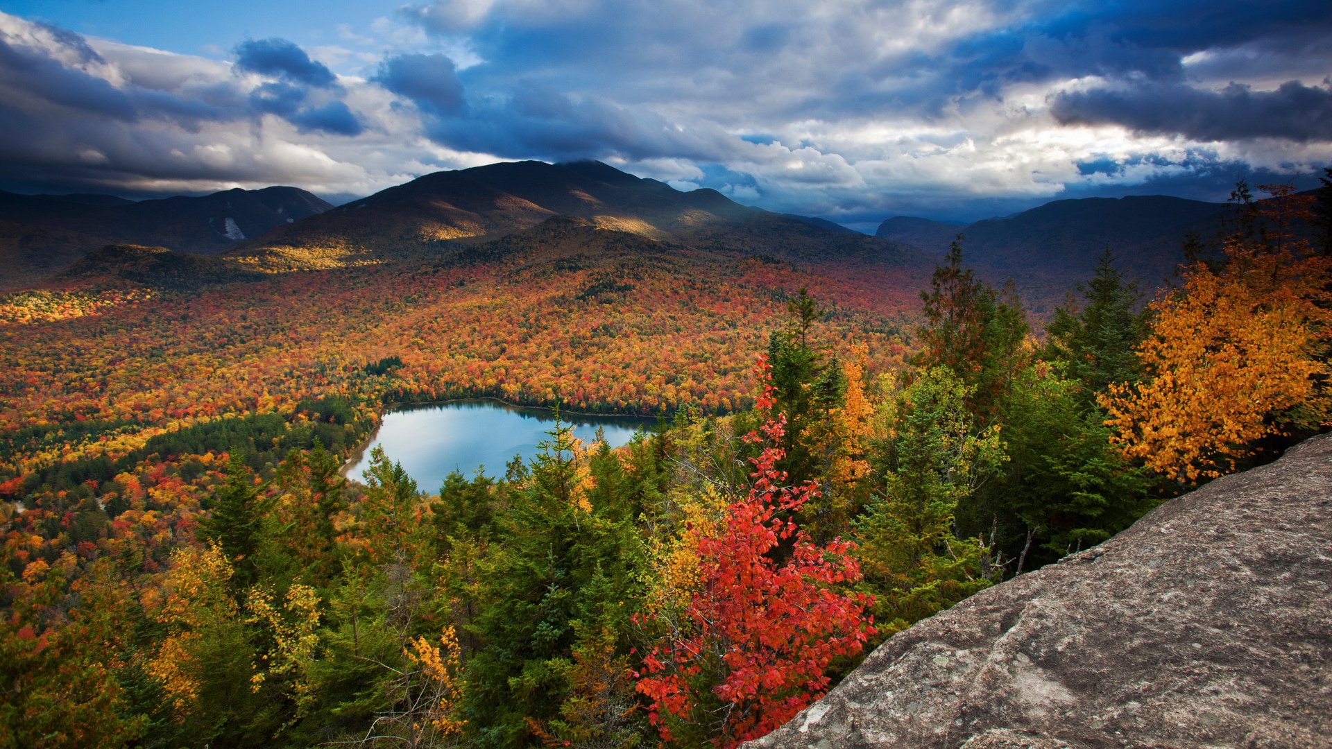 HD desktop wallpaper showcasing a vibrant fall landscape with colorful forest, a tranquil lake, and mountains under a dramatic sky.