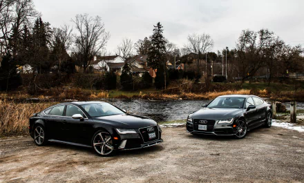 Two black Audi RS7 cars parked near a river with trees and houses in the background, captured in a 4K Ultra HD PC desktop wallpaper image.