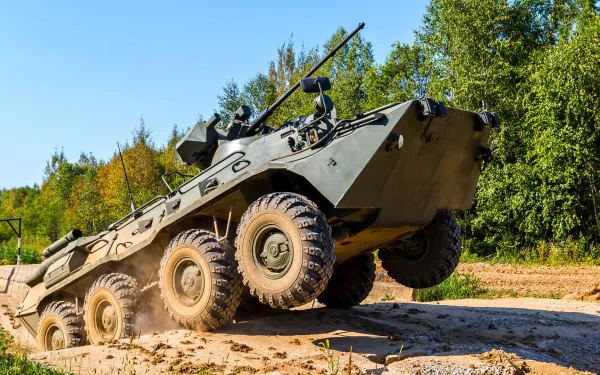 HD desktop wallpaper of a military BTR-80 armored personnel carrier driving off-road against a clear sky and green trees background.