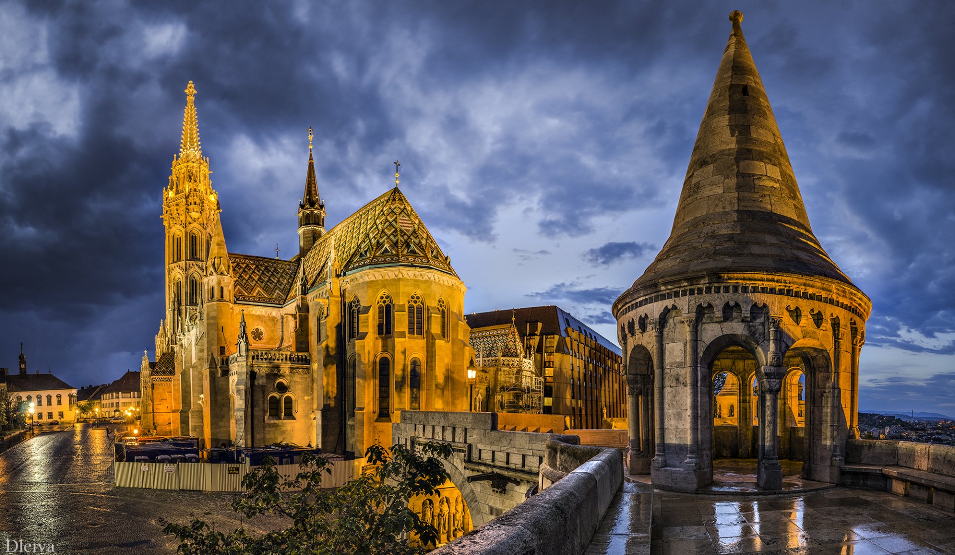 Night view of Matthias Church's intricate architecture and surrounding structures in Budapest, Hungary, beautifully illuminated against a dramatic cloudy sky.