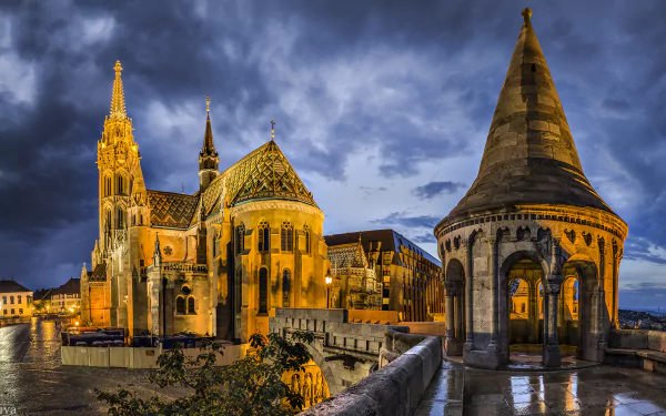 Night view of Matthias Church's intricate architecture and surrounding structures in Budapest, Hungary, beautifully illuminated against a dramatic cloudy sky.