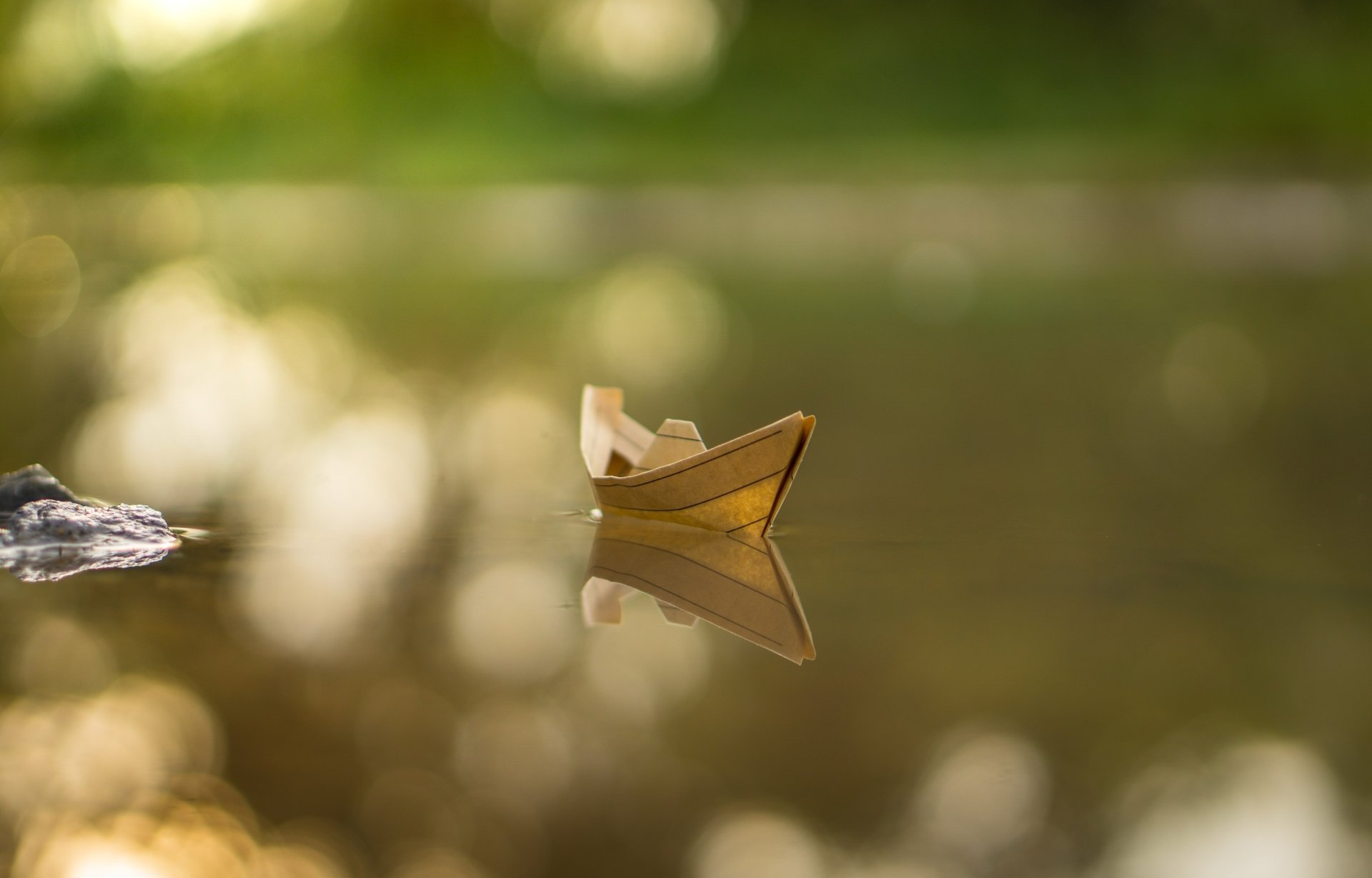 A delicate origami paper boat floats on calm water with a soft bokeh background, creating a serene man-made scene in HD.