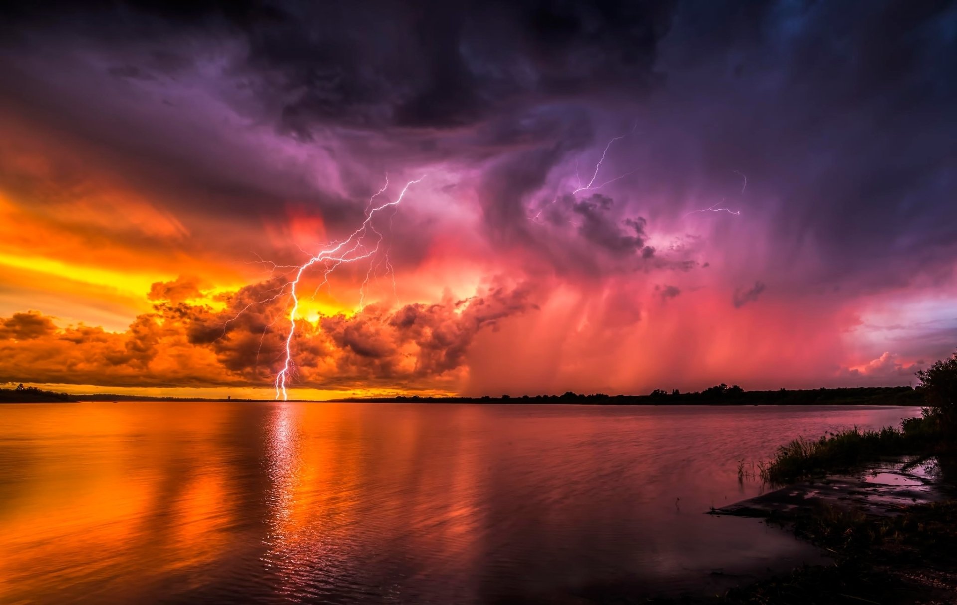 HD desktop wallpaper showing a dramatic sunset over the ocean with storm clouds and lightning illuminating the sky and reflecting on the water's surface.