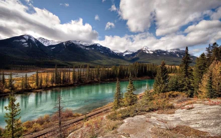 HD desktop wallpaper featuring the stunning Alsek River in Alaska. The image captures the serene river flowing through a lush landscape with majestic mountain peaks in the background under partly cloudy skies.