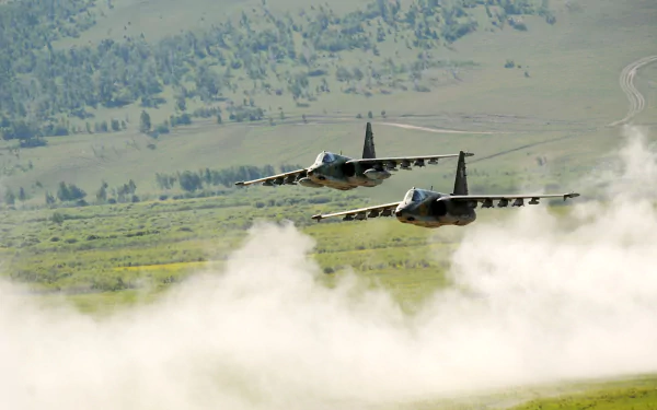 Two Sukhoi Su-25 jet fighters soar through a landscape, creating plumes of dust as they navigate a military exercise. A striking HD wallpaper showcasing powerful aircraft in action.