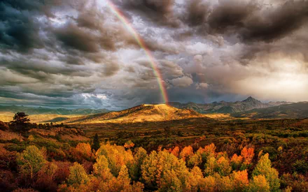 HD desktop wallpaper of a vibrant fall forest landscape under dramatic clouds with a rainbow arching over the colorful autumn trees and distant hills.