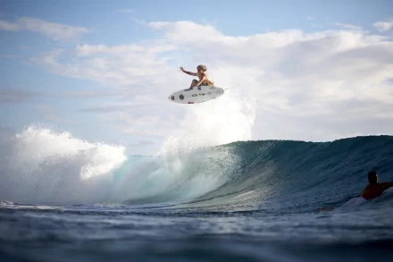 A surfer performs an aerial maneuver over a wave under a partly cloudy sky, captured in an HD desktop wallpaper and background image tagged with surfing and sports.