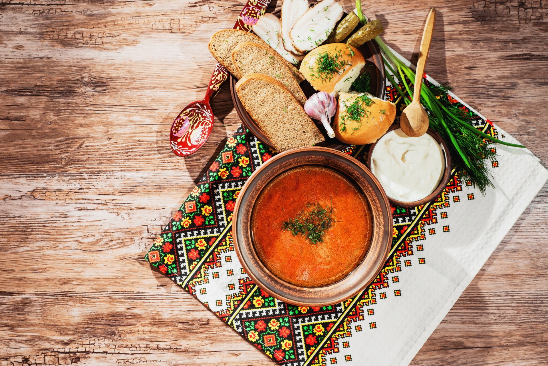 A vibrant still life of garlic soup in a bowl, surrounded by sliced bread, fresh herbs, and garlic cloves on an embroidered cloth, captured in 8K Ultra HD.