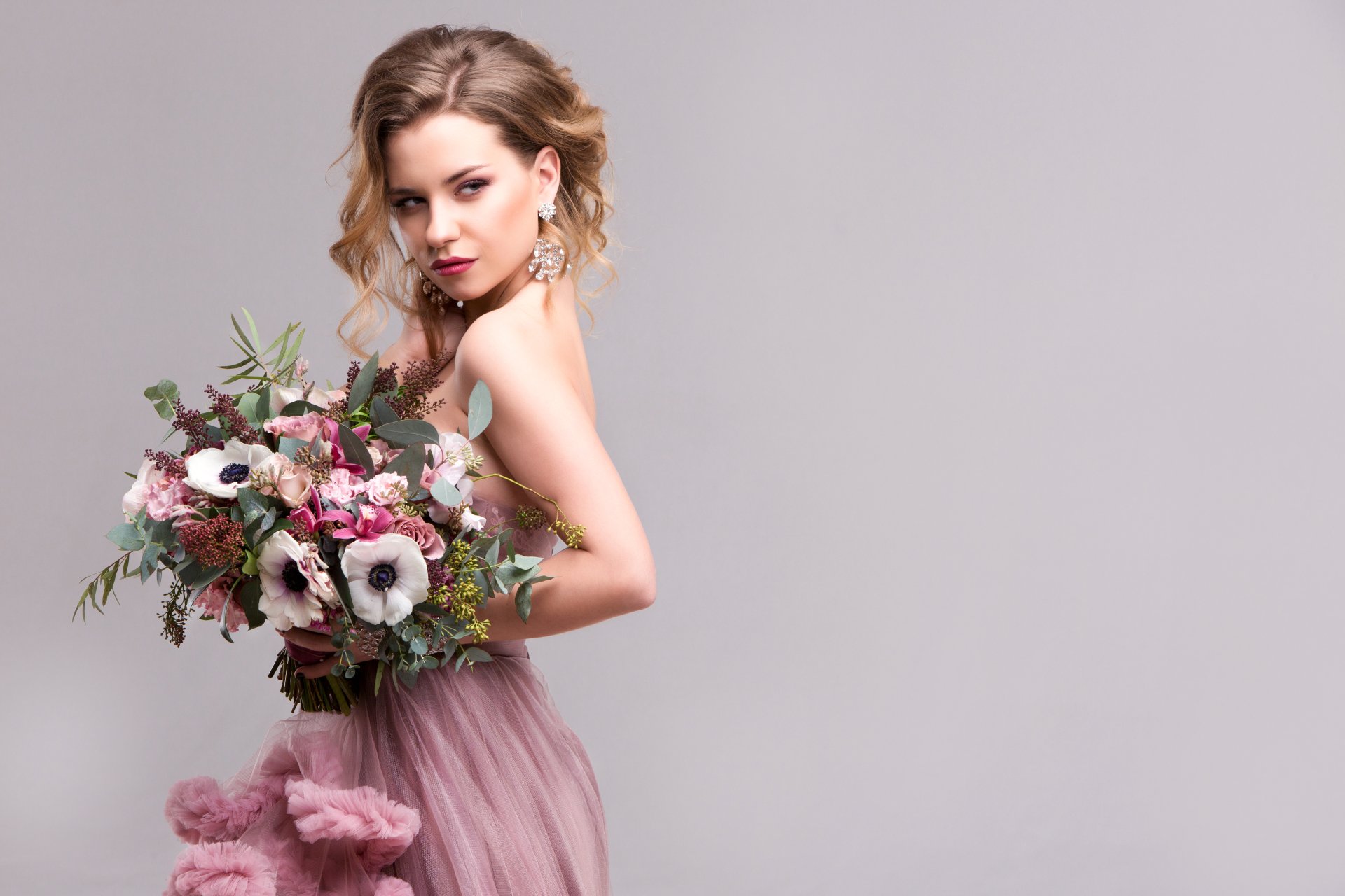 Brunette woman model in a pink dress holds a bouquet of flowers, wearing elegant earrings against a soft grey background in this 4K Ultra HD desktop wallpaper.