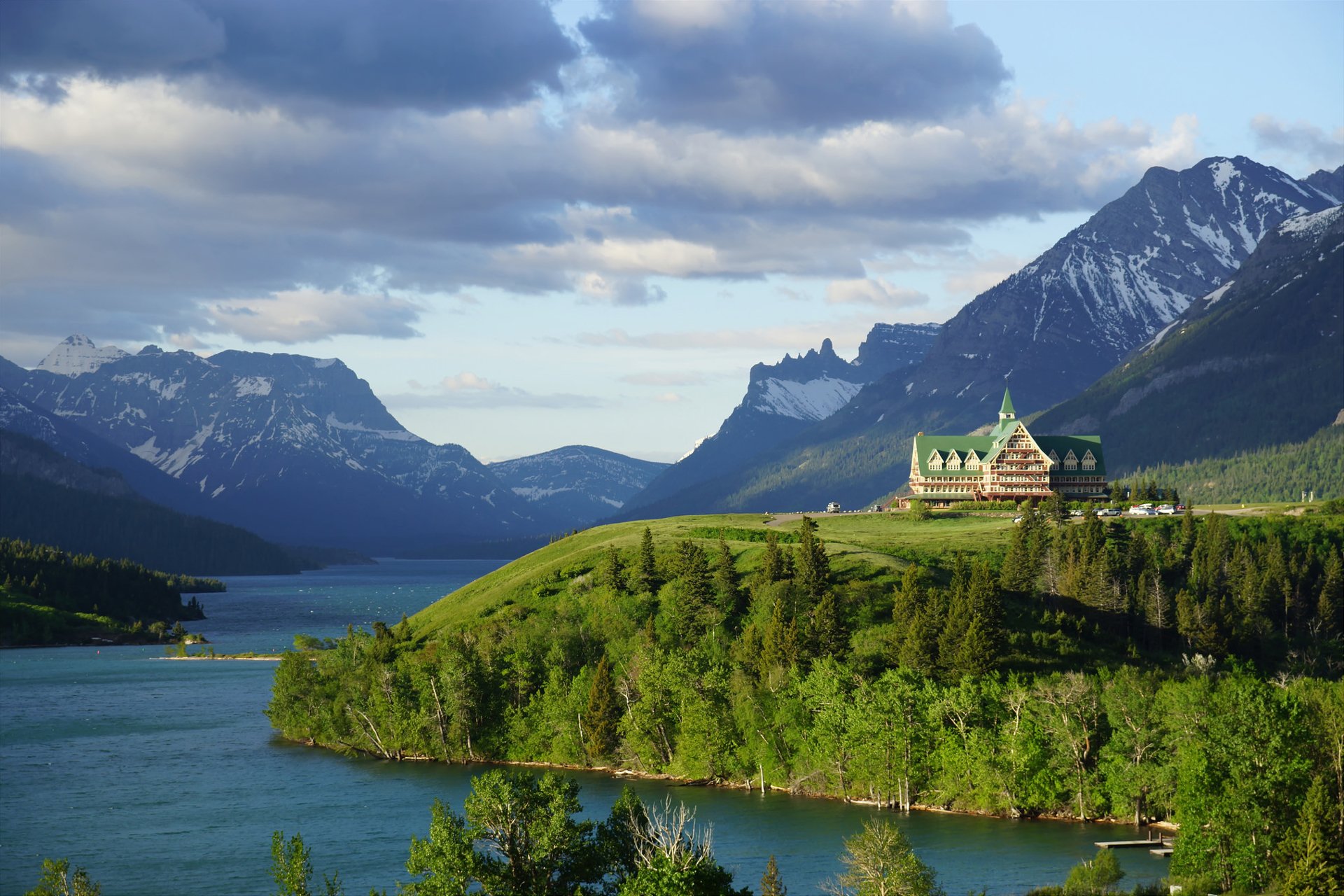 HD PC desktop wallpaper/background: a man-made hotel perched on a forested hill by a blue lake, framed by towering mountains and dramatic cloud-filled sky.