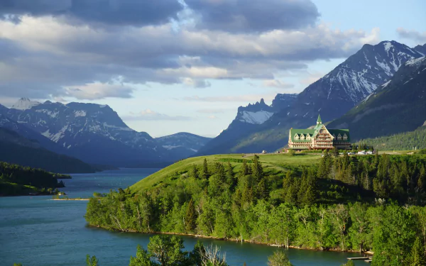 HD PC desktop wallpaper/background: a man-made hotel perched on a forested hill by a blue lake, framed by towering mountains and dramatic cloud-filled sky.