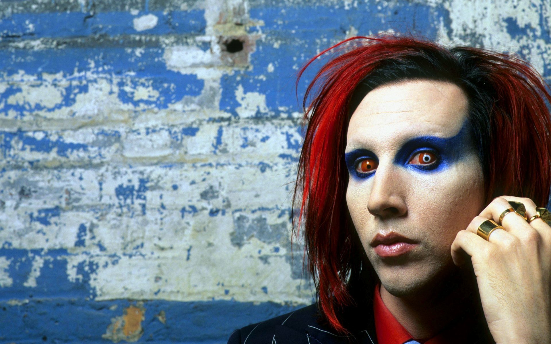 HD PC desktop wallpaper of an industrial/heavy metal musician: red hair, pale skin, bold blue eye makeup, rings on a raised hand and an intense stare against a weathered blue brick wall.