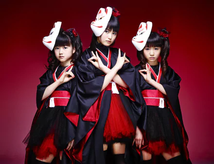 Three Japanese metal idols from Babymetal pose dramatically in traditional attire, showcasing their unique blend of heavy metal and cultural elements against a striking red background.