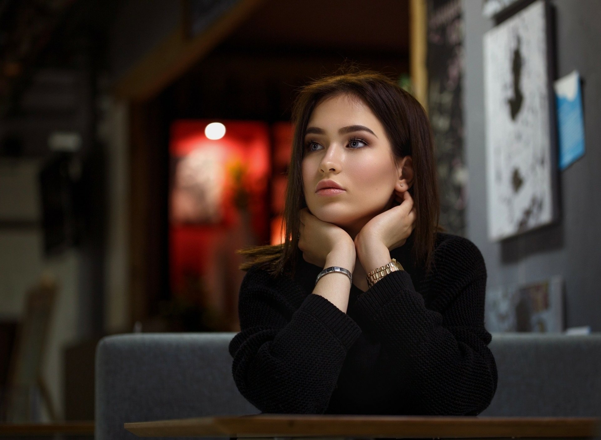 HD desktop wallpaper featuring a brunette model with blue eyes in a contemplative mood, seated indoors against a blurred artistic background.