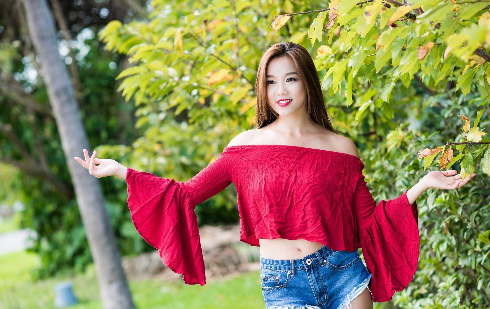 Asian woman with brown eyes and brunette hair, smiling while wearing lipstick, a red off-shoulder top, and denim shorts. She poses outdoors in a natural setting. HD desktop wallpaper and background.