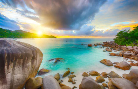 HD PC desktop wallpaper of a Seychelles beach at sunrise, showcasing the ocean, rocky shoreline, lush greenery, and a dramatic sky with clouds over the horizon.